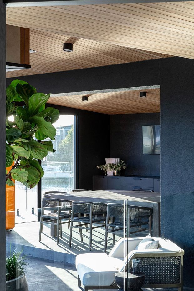 A contemporary open-plan dining and living area with a dark charcoal interior, full-height glazing, and a warm timber-lined ceiling. A rectangular dining table with woven chairs sits behind a white upholstered lounge chair, while indoor planting and a low dark built-in cabinet add texture. Strong daylight enters through the glazing, with an exterior waterfront view visible beyond.