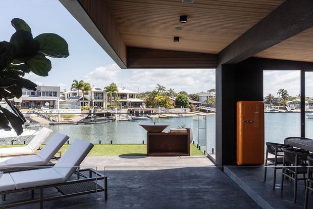 A contemporary covered outdoor living area overlooking a calm waterfront canal or marina. The space features dark concrete flooring, a timber-lined soffit with recessed lighting, substantial dark structural beams, lounge chairs, an outdoor dining setting, a built-in or freestanding bar cabinet, and a bright orange retro-style refrigerator. Water, boats, palms, and neighbouring waterfront homes are visible beyond the open edge.