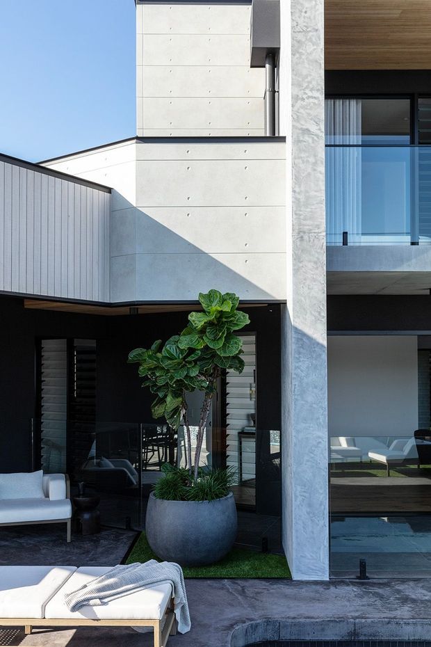 A contemporary multi-level residential exterior with a covered outdoor living area and pool edge in the foreground. The facade combines pale concrete panels with visible form-tie marks, dark framing, expansive glazing, and a vertically ribbed metal-clad upper volume. Outdoor lounge furniture, a large potted plant, timber soffit lining, and a small section of interior living space are visible through the glazing.