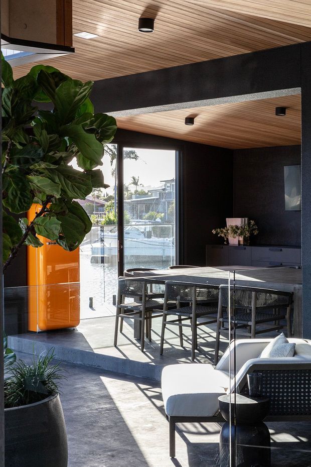 A sophisticated open-plan dining and living area with a dark, dramatic interior palette. The room features a large dining table with woven dining chairs, a contemporary lounge chair and ottoman, dark built-in cabinetry, and a vivid orange cylindrical planter. Warm timber slat ceilings contrast with charcoal-black walls and concrete flooring. Full-height glazing opens toward a marina or waterfront setting, while abundant indoor greenery adds an organic element.
