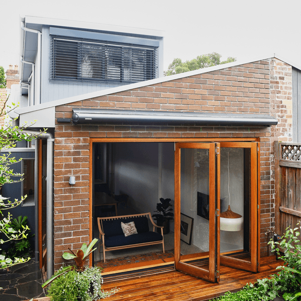 Exterior view of a compact brick extension with a steeply sloping roofline, large timber-framed glazed doors, and a small timber deck opening onto a planted garden courtyard. A taller modern rendered structure with external blinds is visible behind it.