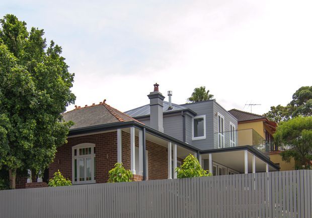 Exterior view of a compact urban residential property, showing a contemporary dark-grey upper-level extension attached to an older red-brick house. The original dwelling has a hipped tiled roof, chimney, and covered front veranda, while the addition features dark cladding, a low-pitched roof, projecting box form, and louvred window screens. A tall grey vertical-slat fence encloses the foreground.