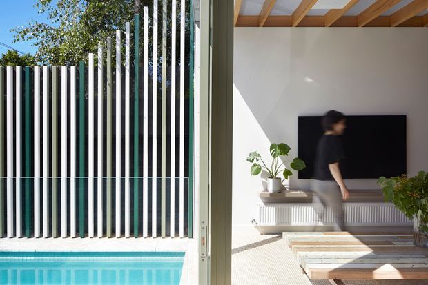 Split architectural view showing a turquoise swimming pool beside a sculptural vertical slatted screen and, through wide open glazing, a bright contemporary living room. The interior features a white wall, wall-mounted black television, long built-in white cabinetry or radiator cover, timber ceiling beams, and a low rustic timber coffee table. Strong sunlight creates a clean indoor-outdoor connection.