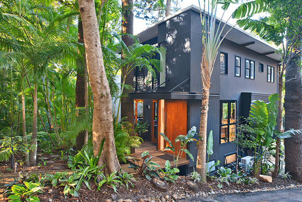 Exterior view of a compact contemporary two-storey house nestled within lush tropical landscaping. The dark charcoal cladding, sharply defined roofline, large windows, timber entry door, and raised timber deck create a modern retreat surrounded by palms and dense greenery.
