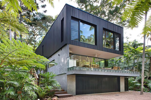 A contemporary multi-storey residence set within lush tropical vegetation. The house features dark vertical metal cladding on the upper level, exposed grey concrete walls below, extensive glazing, a projecting upper storey, a glass-balustraded terrace, and a wide dark garage door at ground level.