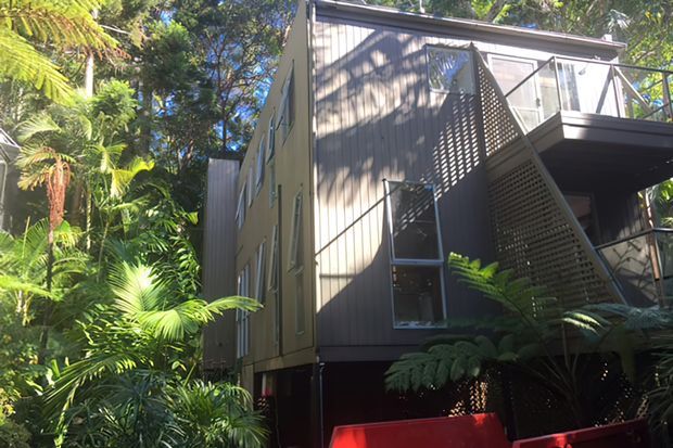 Exterior view of a contemporary multi-level dwelling nestled among dense tropical and subtropical vegetation. The building features dark vertical cladding, extensive glazing, projecting upper-level balconies, and a distinctive perforated privacy screen with strong geometric shadow patterns. The elevated construction and lush setting create a secluded, forested residential character.
