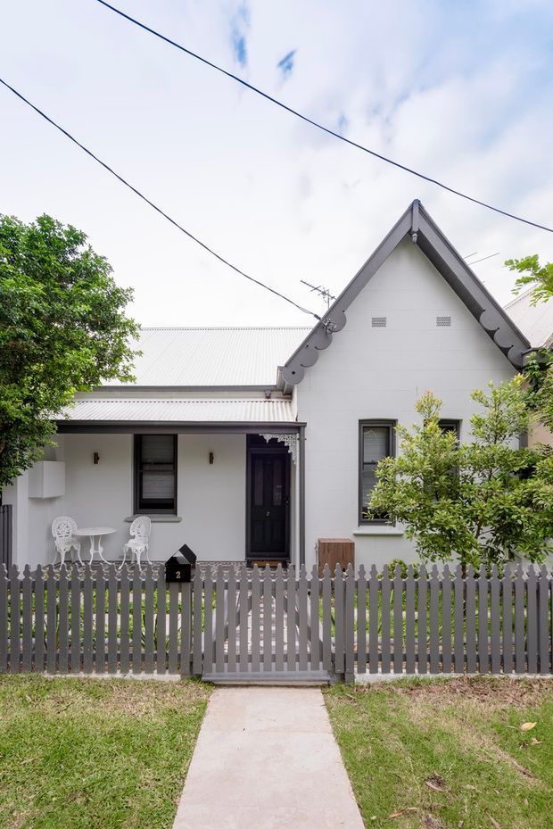 Front exterior view of a compact heritage-style cottage behind a grey picket fence. The white-painted facade has a steep gabled roof with decorative scalloped verge detailing, corrugated metal roofing, dark-trimmed windows and a covered front porch with small outdoor seating. A concrete path leads through the front lawn to the entry.