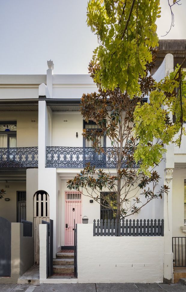 Street-facing view of a narrow, multi-storey terrace or townhouse facade with white-painted masonry, ornate dark metal balcony balustrades, a small front courtyard enclosed by a picket fence, and a distinctive pink front door. Mature planting partially screens the facade, while a covered upper-level balcony and traditional decorative details are visible.