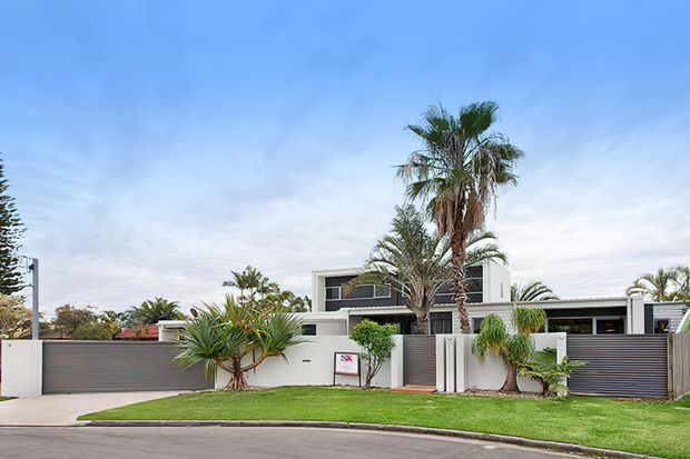 Street-facing view of a contemporary two-storey residence with a white rendered facade, broad horizontal dark-grey fencing and garage doors, flat rooflines, and tropical palm landscaping. The property has a low, strongly horizontal composition and a manicured front lawn.