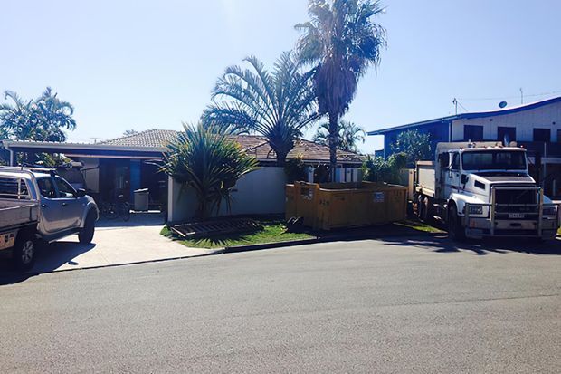 Street-level exterior view of a low-rise suburban property with a tiled-roof dwelling, a blue flat-roof commercial or industrial-looking building, palm landscaping, paved frontage, and parked utility vehicles. The image is dominated by the building exteriors and streetscape.
