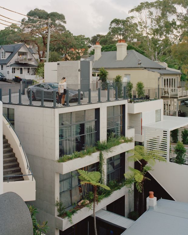 Elevated exterior view of a contemporary multi-level residential development built into a sloping, leafy suburban site. The foreground building has a raw concrete facade, large black-framed glazed openings, planted terraces and an external stair, with a white louvred privacy screen and covered outdoor areas. Older pitched-roof homes with chimneys are visible around it, along with fencing, parked cars and mature trees.