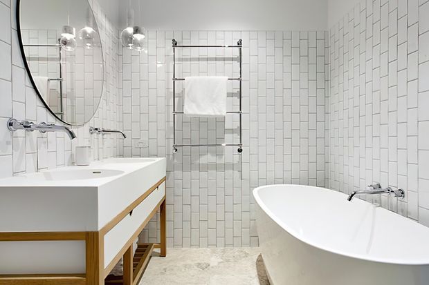 Bright contemporary bathroom featuring white vertically stacked rectangular wall tiles, a freestanding white bathtub, a floating white vanity with timber trim, a large round mirror, chrome fixtures, and a wall-mounted heated towel rail. The room has a clean, refined, high-contrast white palette with warm timber accents.