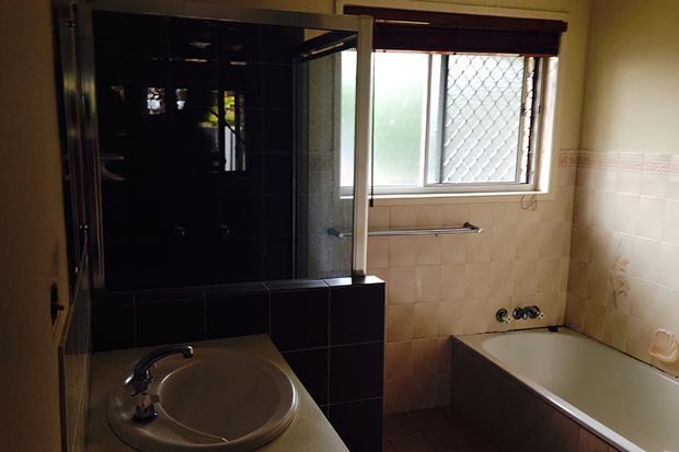 A compact, older-style bathroom with a white corner basin and chrome tap in the foreground, a white bathtub along the right wall, and a dark tiled shower enclosure at the rear. Beige wall tiles with a decorative border, a frosted window with security mesh, and simple chrome fixtures complete the room.