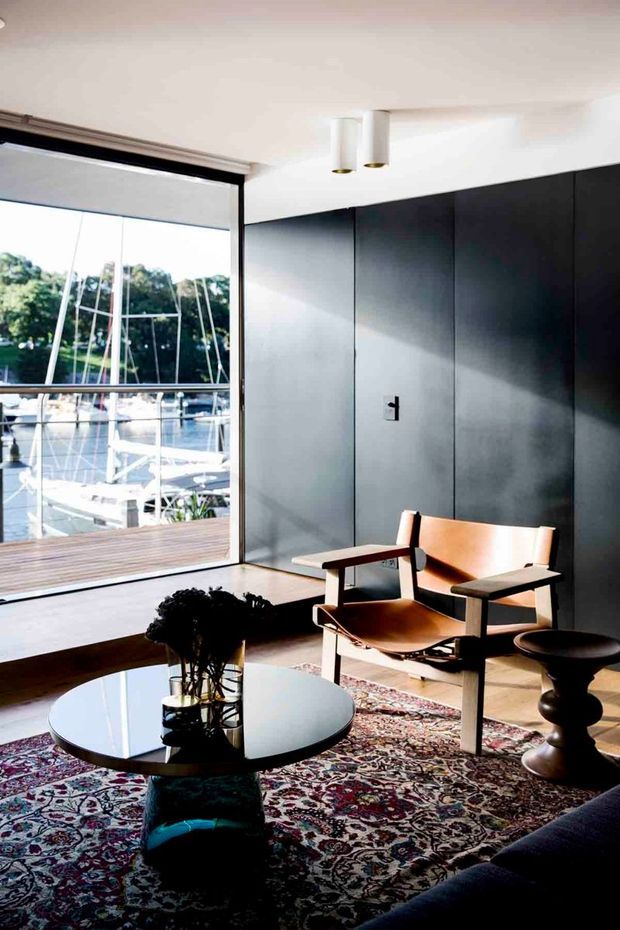 A refined modern living room with a large glazed opening overlooking a marina, dark charcoal built-in wall panelling, white ceiling, timber flooring, and a patterned Persian-style rug. A low round black reflective coffee table, sculptural side table, and leather-and-timber lounge chairs create a warm mid-century-inspired seating area. Strong daylight enters from the waterfront-facing glazing.