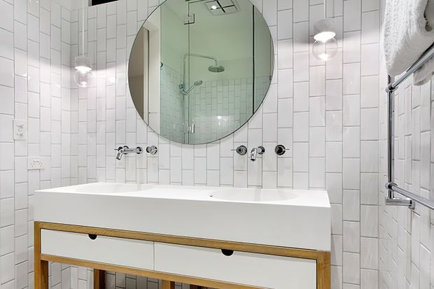 Bright contemporary bathroom featuring a wall-mounted double vanity with white drawers and a timber-toned open-frame base, white vertically arranged rectangular wall tiles, chrome tapware, integrated lighting, and a large round mirror reflecting a glass shower enclosure.