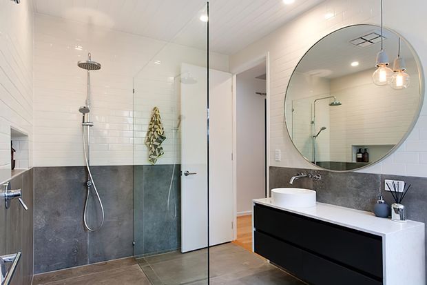 Bright contemporary bathroom with a walk-in glass shower, white subway-tile walls, dark grey tiled lower walls, and a floating black vanity with a white vessel basin. A large round mirror and pendant lights add a refined focal point, while timber flooring is visible through the open doorway.