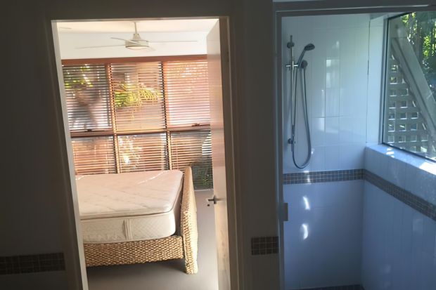 Interior view showing a tiled bathroom in the foreground/right with a walk-in shower, handheld shower rail, glass enclosure, and a horizontal dark mosaic tile border. Through the adjacent opening is a bedroom with a woven bed base, mattress, timber venetian blinds, and ceiling fan. Natural light enters through the bathroom window and bedroom glazing.
