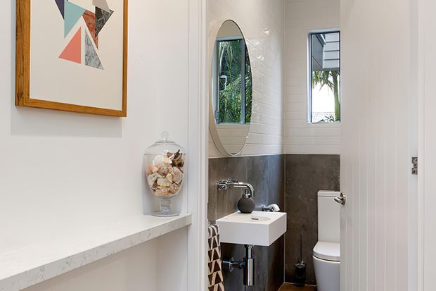 Compact contemporary bathroom viewed from an adjacent hallway or entry area. The room features a wall-mounted white basin with chrome tapware, a white toilet, dark grey tiled lower walls, white vertical wall tiles, a narrow window, and an oval mirror. A white stone-look shelf and framed abstract artwork are visible outside the bathroom.