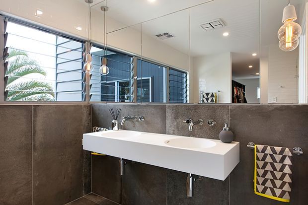 Contemporary bathroom with a floating double vanity in white, dark grey tiled walls and floor, wall-to-wall mirror, chrome tapware, and pendant lighting. A louvred window provides views to palm foliage and an exterior blue-clad structure.