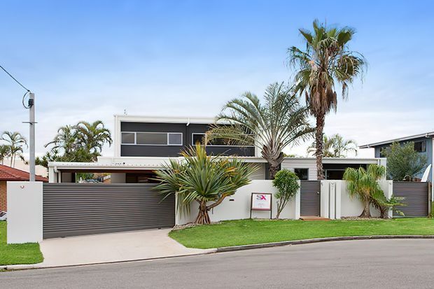 Street-facing view of a contemporary two-storey residence with a flat roof, dark horizontal slatted fencing and garage door, white rendered walls, broad glazing, and a landscaped front garden featuring palms and sculptural tropical planting.