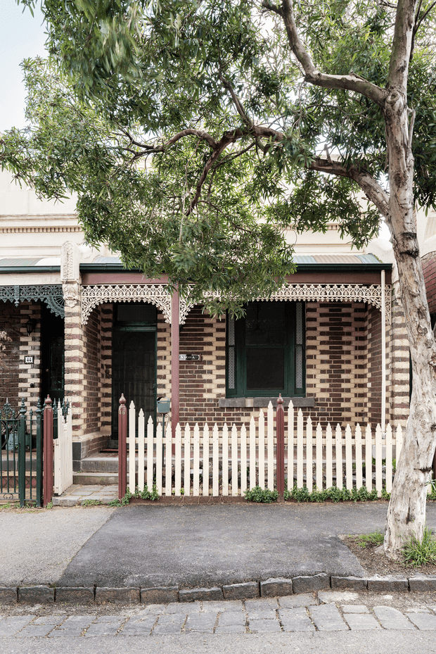 Street-facing view of a heritage terrace-style brick cottage with distinctive striped brickwork, ornate lacework verandah detailing, dark green timber-framed windows and doors, a white picket fence, and a mature street tree. The image primarily captures the residential facade and front boundary from the pavement.