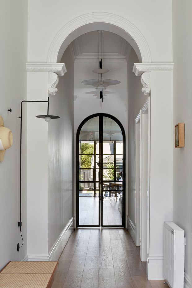 A refined residential hallway with a long light timber floor, white decorative wall mouldings and skirting, and a dramatic arched opening with ornate classical detailing. A black-framed arched glass door leads to a bright outdoor or courtyard area. Slim black wall lights and sculptural pendant lights accent the corridor, while a woven bench is partially visible in the foreground.