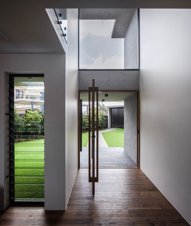 A modern residential entry hall with dark timber flooring, white walls, and a tall glazed pivot door opening to a landscaped courtyard. A large overhead skylight or clerestory brings daylight into the narrow hallway, while a louvred side window provides views toward the garden and neighbouring residences. Concrete and timber elements create a clean, restrained architectural composition.