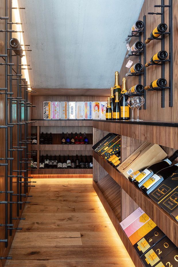 A narrow, carefully fitted wine cellar or wine display room with floor-to-ceiling timber cabinetry and shelving. Bottles are stored in illuminated open shelves and black metal wall racks, with champagne bottles and wine glasses displayed on a central counter. The room has warm timber flooring and joinery, a textured pale ceiling or wall surface, integrated warm LED lighting, and a refined retail-like presentation.