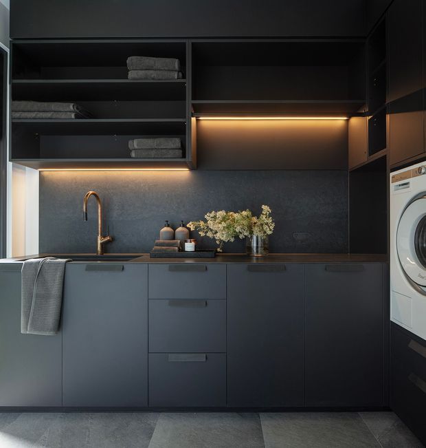 A refined dark-toned laundry room with matte charcoal cabinetry, open shelving holding folded towels, an integrated sink with a warm brass tap, a dark textured splashback, under-cabinet lighting, and a front-loading washing machine concealed within the joinery. Grey floor tiles and warm accent lighting create a moody, luxurious atmosphere.