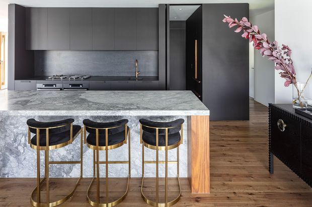A refined contemporary kitchen with a large veined stone island, three black upholstered bar stools with brass frames, matte dark cabinetry, integrated appliances, and a dark stone backsplash. Warm timber flooring and timber island detailing add contrast, while a tall dark doorway and decorative floral branch are visible in the background.