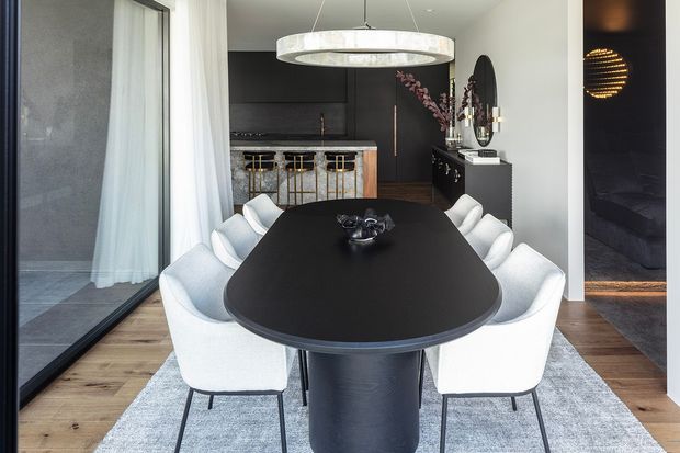 Contemporary dining area with a large black oval dining table and white upholstered chairs, positioned on a pale rug over timber flooring. A dark, sophisticated kitchen with integrated cabinetry and a marble-look island is visible behind, while a sculptural ring pendant light hangs above the table. Full-height glazing with sheer curtains and a dark-toned adjoining room reinforce the refined monochromatic palette.