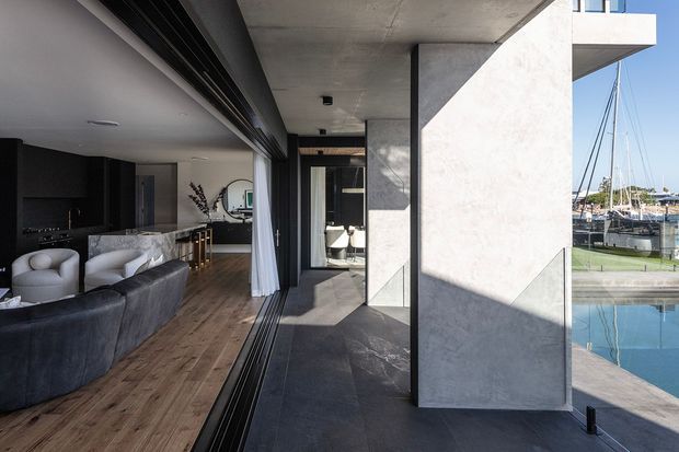A contemporary waterfront apartment or home interior opening onto a covered terrace beside a swimming pool. The open-plan living area includes dark built-in cabinetry, a pale stone kitchen island, curved light armchairs, a dark sofa, timber flooring and full-height sliding glazing. The terrace has dark tiled flooring, pale concrete or rendered walls and strong geometric sunlight and shadows, with marina boats visible beyond.