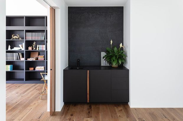 A refined built-in storage and media cabinet occupies a recessed wall, with matte black cabinetry, a dark textured feature wall, and a slim timber handle detail. A tall indoor plant and framed artwork sit on the cabinet. Through the open doorway, a built-in bookshelf and part of an adjacent study or living area are visible. Warm timber flooring contrasts with the monochrome joinery and white walls.