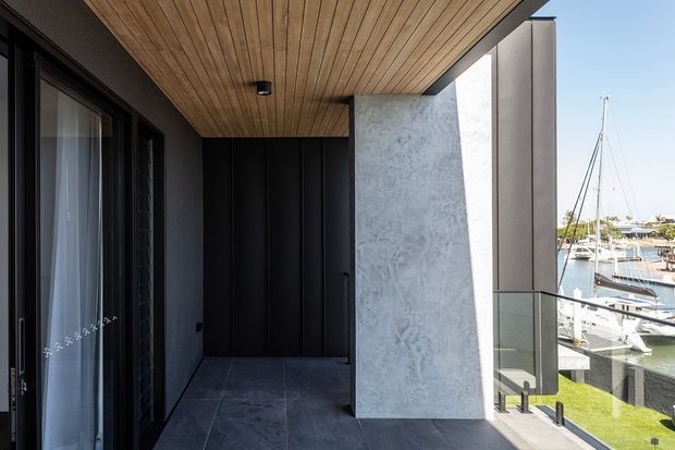 A contemporary covered balcony or terrace viewed from beside a dark-framed glazed opening. The space features large dark grey floor tiles, black vertical wall cladding, a timber-lined soffit with recessed lighting, and a prominent light grey textured concrete or stone privacy wall. Glass balustrading overlooks a marina with boats and waterfront buildings.