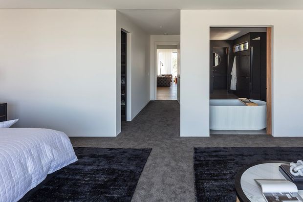 A contemporary bedroom viewed toward a central hallway, with a partially visible bed at left, dark textured area rugs over charcoal carpet, white walls, and a built-in bathroom or dressing area opening on the right. The room has clean-lined recessed architecture, dark timber or charcoal joinery, and a calm monochromatic palette.