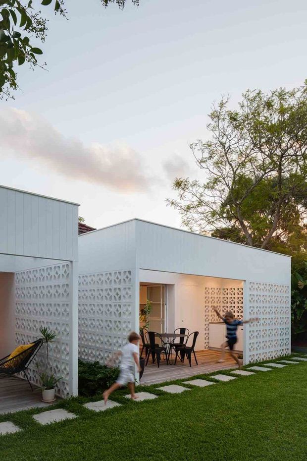 Exterior view of a low, white modern pavilion or house set within a landscaped garden. The building has crisp flat rooflines, white vertical cladding, and extensive decorative breeze-block screens enclosing a covered timber deck or patio. A small outdoor dining setting is visible beneath the covered area, with stepping-stone paths through the lawn and mature trees surrounding the structure.
