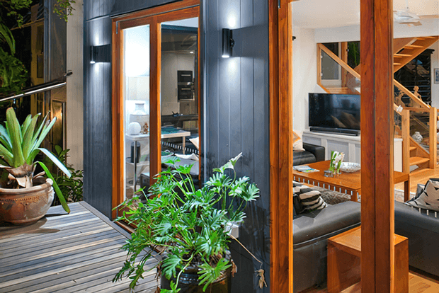 A contemporary residential exterior and deck area viewed through lush planting, featuring dark vertical cladding, warm timber-framed glazing and a timber-framed entry. The interior beyond the glazing includes a living room with dark seating, a television, timber furniture and a visible timber stair structure.