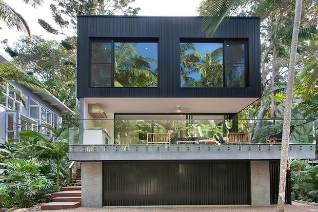 Contemporary two-storey residence set among lush tropical vegetation, with a strongly cantilevered upper volume clad in dark vertical metal, expansive glazed openings, a transparent ground-level living area, and a broad glass-balustraded terrace above an integrated garage.