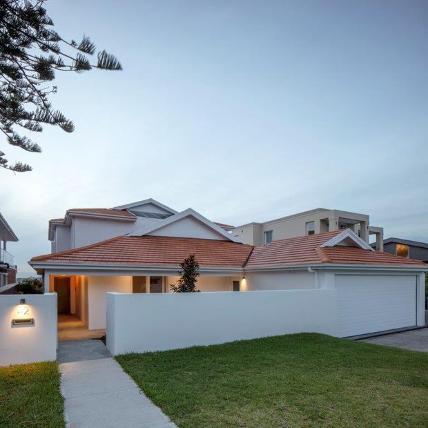 Street-facing exterior view of a contemporary suburban residence at dusk, featuring white rendered walls, multiple terracotta-tiled pitched roofs, a large integrated garage, covered entry, front lawn, and concrete pedestrian path. A neighbouring modern multi-storey building and coastal-style tree are visible in the background.