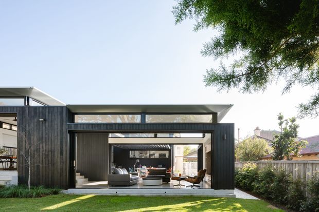 Exterior view of a contemporary single-storey house extension with dark vertical cladding, broad open-plan glazing and sliding doors, and strong horizontal roof planes. The central opening reveals a living room inside, while a lawn, boundary fencing, and mature trees frame the property.