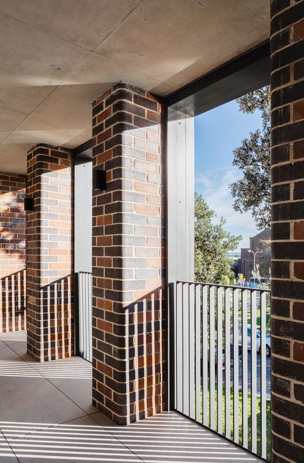 A covered exterior balcony or walkway with robust dark red and grey brick columns, a raw concrete soffit, black metal-framed glazing, and vertical metal balustrades. Strong sunlight casts striped shadows across the tiled floor, while trees, neighbouring buildings, and a street are visible beyond the railing. The scene has a sturdy, contemporary institutional or apartment-building character.