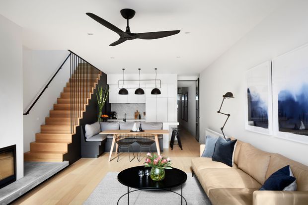 Bright open-plan living and dining area with a compact kitchen at the rear. The room features a timber staircase with black metal balustrade, pale timber flooring, white walls and cabinetry, a large beige sectional sofa, black round coffee table, contemporary pendant lights, ceiling fan, and abstract blue artwork. The space has a clean, contemporary residential interior with strong black-and-white contrast and warm timber accents.