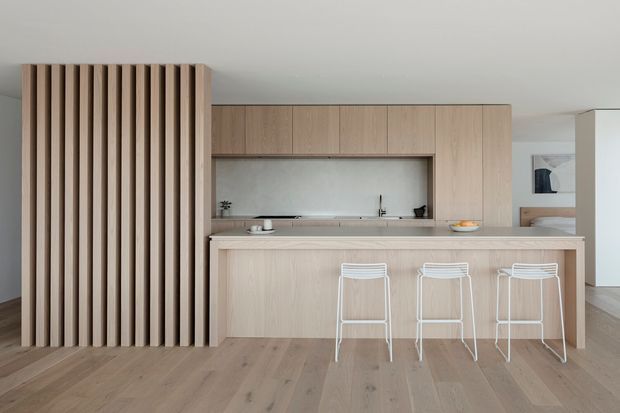 Minimal contemporary kitchen with a long pale timber island and three white wire-frame stools. Full-height light timber cabinetry surrounds a recessed backsplash and sink, while a vertical timber slatted screen partially conceals the kitchen at left. Wide timber floorboards and a restrained neutral palette create a clean, calm composition.