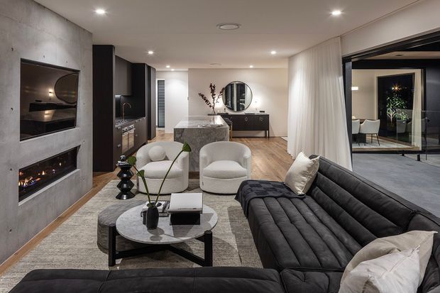 Contemporary open-plan living area with a large black sectional sofa, round coffee table, sculptural lounge chairs, and a textured rug. A concrete feature wall with built-in fireplace openings anchors the room, while a dark kitchen with a stone island sits behind the living zone. Floor-to-ceiling curtains and black-framed glazed doors connect to an adjacent dining area or courtyard.