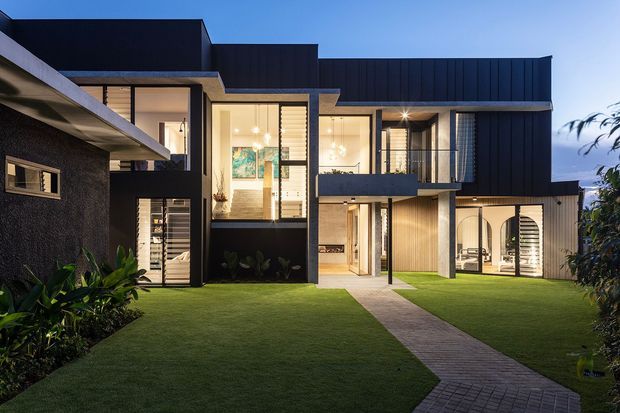 Exterior dusk view of a contemporary multi-level residence with strong geometric massing, dark vertical cladding, pale rendered or concrete elements, extensive floor-to-ceiling glazing, balconies, and a landscaped lawn. A paved path leads through the front garden to the illuminated entrance and living spaces.