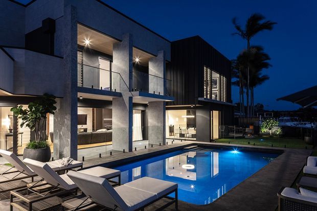 Night-time exterior view of a contemporary multi-level residence with a rectilinear concrete and dark-clad facade, extensive glazing, balconies, and warm interior lighting. A blue swimming pool, paved pool terrace, outdoor lounge furniture, umbrellas, lawn, and palm trees occupy the foreground and side yard.