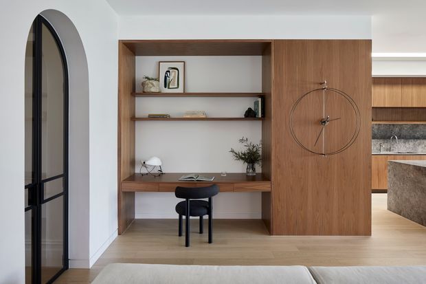 A refined open-plan interior featuring a built-in timber study nook and shelving integrated into a large timber wall unit. The compact desk has a black chair, books, artwork and a small plant, while a matching timber panel incorporates a minimalist wall clock. An arched black-framed glazed doorway is visible to the left, and part of a contemporary kitchen with timber cabinetry, stone surfaces and a peninsula appears to the right. The foreground includes the edge of a sofa or bed.