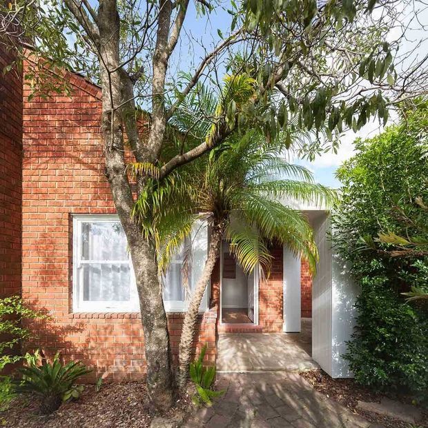 Exterior view of a red brick residential building partially screened by mature trees and palms. A white-framed window and recessed entry porch with a contrasting white wall are visible, approached by a paved walkway through landscaped planting.