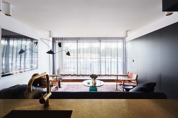 A contemporary open-plan living room viewed from behind a kitchen or bar counter. The space features a dark sofa in the foreground, mid-century-style timber and leather lounge chairs, a glass coffee table, a patterned area rug, sculptural black floor lighting, full-height sheer curtains, and a large dark built-in wall or cabinetry element. A brass-toned kitchen tap and dark sink are visible in the foreground, while framed artwork or a reflective glazed opening occupies the left wall.