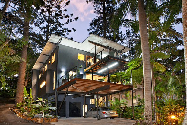 A multi-level contemporary house nestled in dense tropical vegetation. The dark-clad structure features projecting roof planes, extensive glazing, open balconies, timber-lined decks, and a covered carport beneath the elevated living spaces.